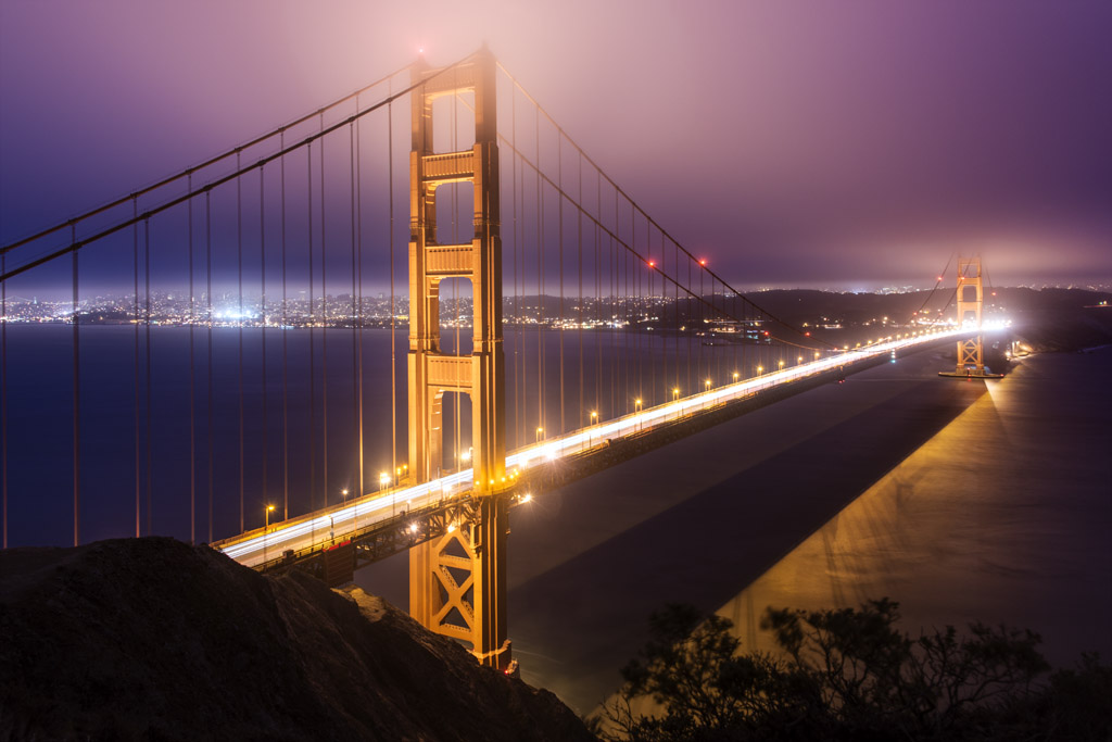 This photo is a long exposure of the Golden Gate Bridge, San Francisco Bay, California.  Lights from the cars trace light trails along the deck of the bridge.  Lights on top of the bridge's towers light up mist above the bridge creating a soft glow in the sky above.