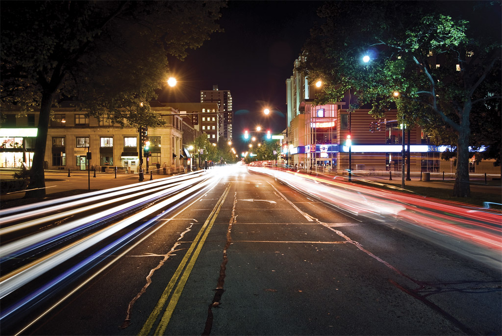 A photograph looking down Green Steet towards Campus Town near the University of Illinois at Urbana-Champaign (UIUC).  Light trails created by the lights of cars passing hover just above the road's surface.  Those light trails, street lights, traffic lights, and various buildings light up the scene.